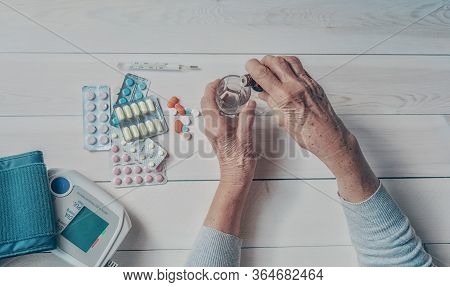 Senior Hands With Medicine Bottle Putting Drops In A Glass, Pills, Tonometer, Thermometer On A Table