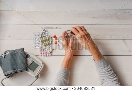 Senior Hands With Medicine Bottle Putting Drops In A Glass, Pills, Tonometer, Thermometer On A Table
