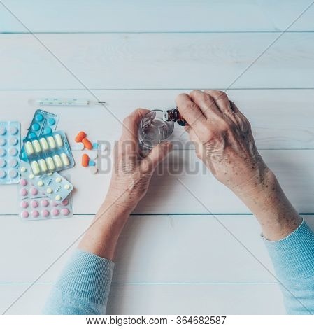 Senior Hands With Medicine Bottle Putting Drops In A Glass, Pills, Tonometer, Thermometer On A Table