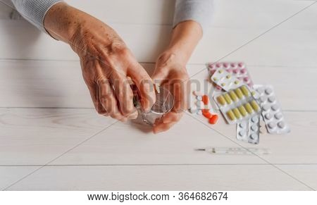 Senior Hands With Medicine Bottle Putting Drops In A Glass, Pills, Tonometer, Thermometer On A Table