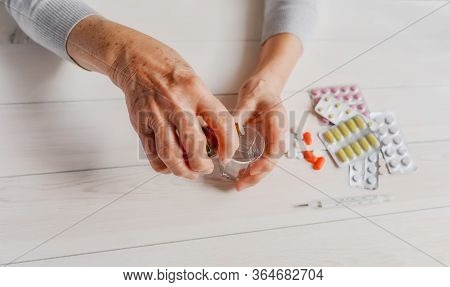 Senior Hands With Medicine Bottle Putting Drops In A Glass, Pills, Tonometer, Thermometer On A Table