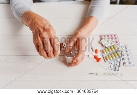 Senior Hands With Medicine Bottle Putting Drops In A Glass, Pills, Tonometer, Thermometer On A Table