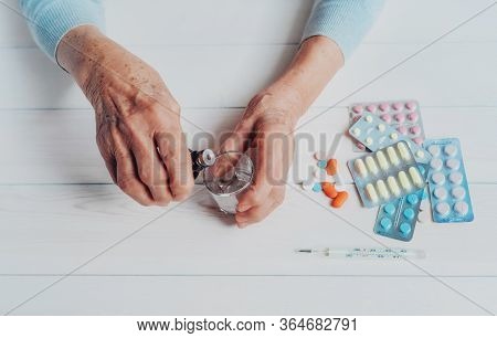 Senior Hands With Medicine Bottle Putting Drops In A Glass, Pills, Tonometer, Thermometer On A Table