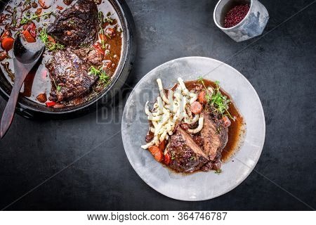 Traditional German braised beef cheeks in brown red wine sauce with noodles and carrots offered as top view on a modern design plate and stewpot on an old rustic board 