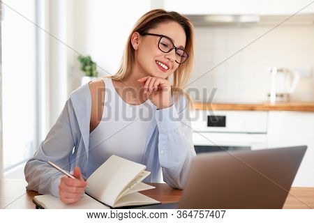 Smiling Woman Working On Laptop At Home On Kitchen. Writing On Notepad. Business Woman Thinking Abou
