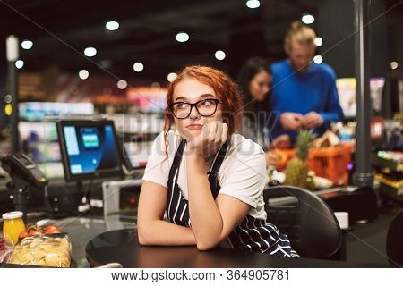 Young Boring Cashier In Eyeglasses And Striped Apron Dreamily Looking Aside Working In Modern Superm