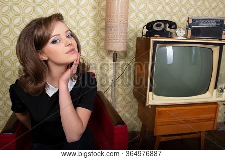 Young smiling ecstatic woman looking at camera and happy smiling in room with vintage wallpaper and interior, retro stylization 60-70s. Furniture, tv set and another technique of the mid 20th century
