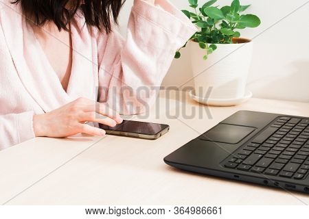 Woman In Bathrobe Sitting At The Table At Home With Laptop And Phone. Remotely Work At Home. Distanc