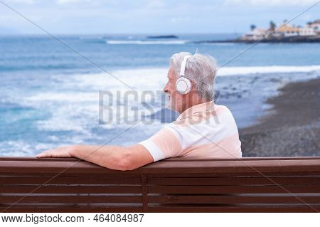 Senior Bearded Man Wearing Headphones Sitting On Outdoors Bench At Sea Looking At Horizon Enjoying F