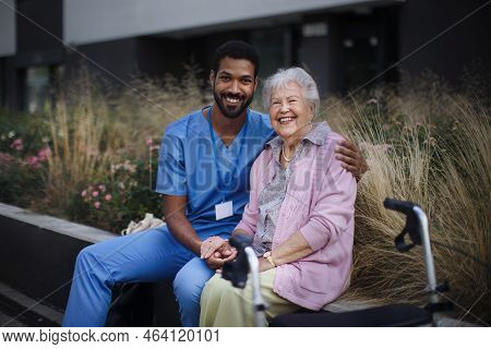 Happy Senior Woman Sitting And Talking With Medical Assistant In Front Of Nurishing Home.