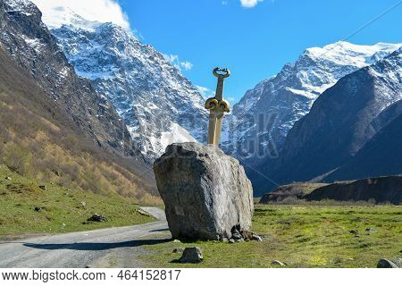 A Sword In A Stone On A Mountain, A Road Passes Nearby, Snow-capped Mountains And A Gorge, Green Gra