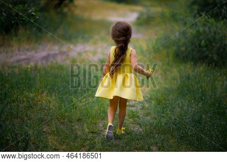 Little Blonde Curly Girl In Yellow Dress And Sneakers Walking And Looking Up Over Grey Wall Backgrou