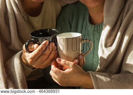 Grandparents Wrapped In Warm Blanket Hold Cups Of Hot Tea Or Coffee, Feeling Cold At Home. Home Heat