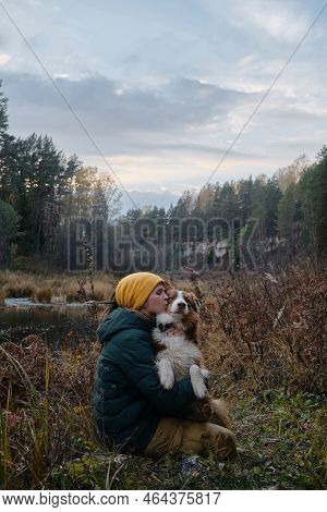 Young Caucasian Woman Sitting Happily With Dog In Autumn Forest By River. Female Pet Owner Hugs And 