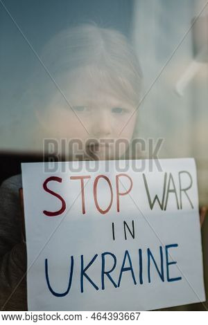 Child At The Window And Calls No War. Poor Girl Protesting War Conflict Raises Banner With Inscripti