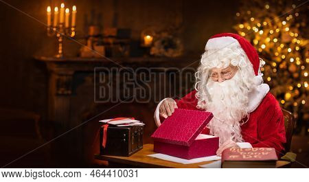 Workplace Of Santa Claus. Cheerful Santa Is Conjuring Over The Gift Box While Sitting At The Table. 