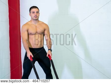 A Handsome Muscular Young Man Using Battle Ropes For Exercise In A Gym. He Focuses On Performing The