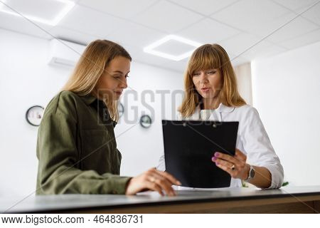 Medical Receptionist Gives Forms To Fill To Female Patient Who  Standing At Reception Counter In Mod