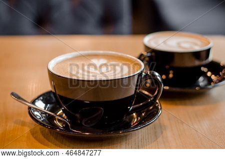 Two Big Black Mugs With Saucer Containing Latte Or Cappuccino Coffee Drink On A Wooden Table In Cafe