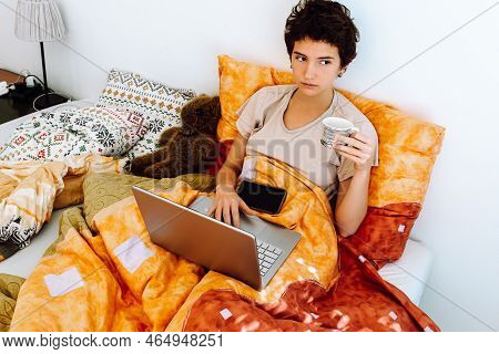 Teenager Sitting In Bed Under Blanket, Holding Coffee Cup, Working Or Studying Online On Laptop With