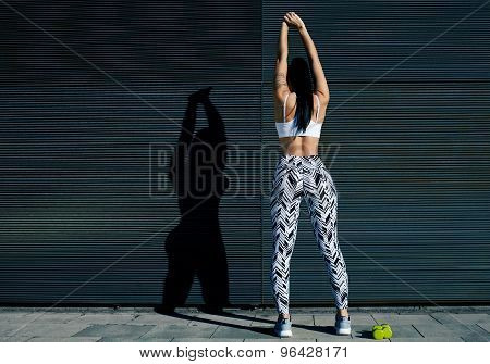 Back view portrait of young woman with perfect figure stretching her hands against black wall