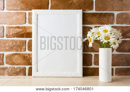White frame mockup with daisy bouquet in styled vase near exposed brick wall. Empty frame mock up for presentation design. Template framing for modern art.