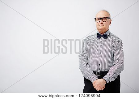 Elegant pensioner in glasses and striped shirt with bow tie standing with clasped hands and looking at camera with serious face expression