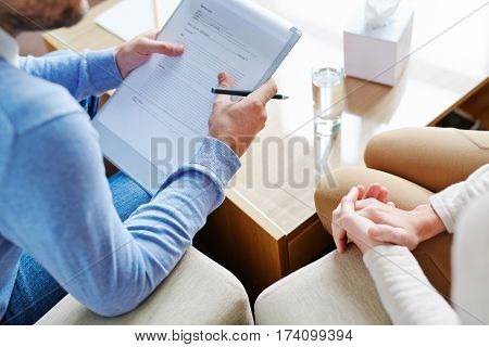 Mature psychiatrist sitting next to his female patient and asking her questions necessary for filling in medical card, close-up shot