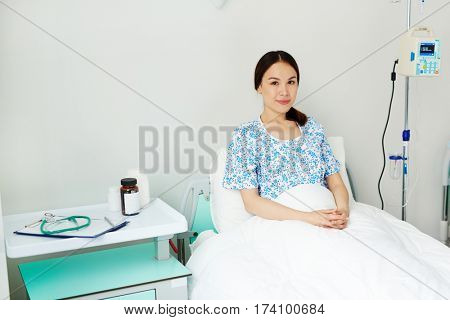 Portrait of young pregnant woman lying in bed in comfortable ward, waiting for gynecological check-up and smiling happily