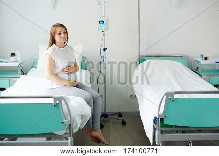 Young pregnant woman sitting on bed in comfortable ward, waiting for doctor and stroking her belly thoughtfully