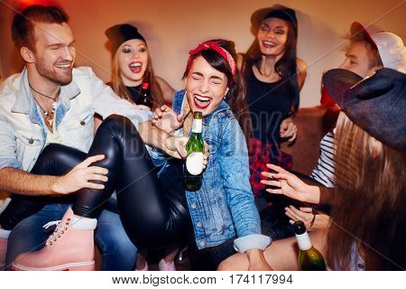 Group of trendy young people getting drunk at late night swag party, going crazy and laughing while sitting on sofa with beer bottles