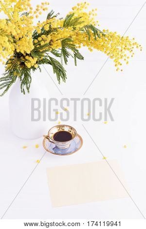Cup of coffee and empty card on light wooden table and blooming mimosa