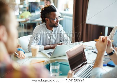 Waist-up portrait of African American employee with dreads looking at his colleague while listening to her with concentration during meeting in boardroom