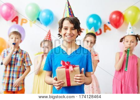 Portrait of excited birthday boy holding box with present standing in front smiling happily with his friends blowing party noisemakers behind in background