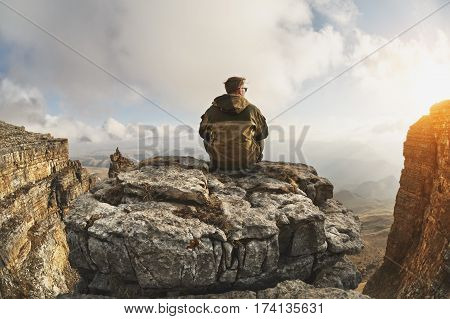 A young man sits on a rock surrounded by cliffs, located above the clouds in a great location, on a background of clouds, valleys, fields, mountains and sunset