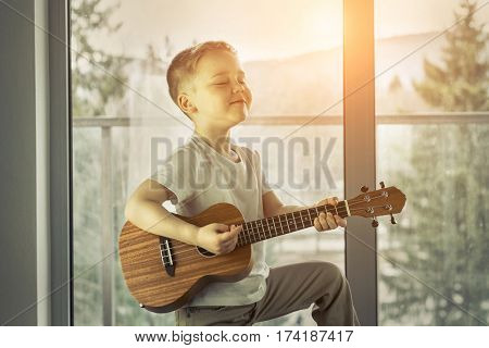 Young boy play on guitar at home at sunny day. Boy play on ukulele - hawaiian guitar.