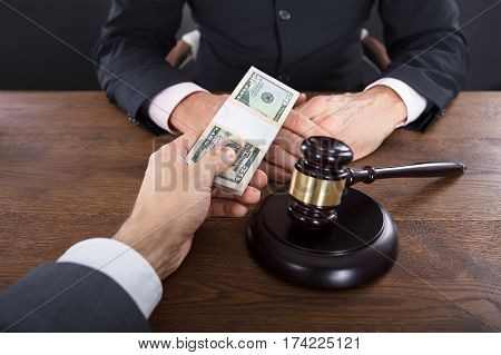 Close-up Of A Businessman Giving A Bribe To A Judge With Gavel On Wooden Desk