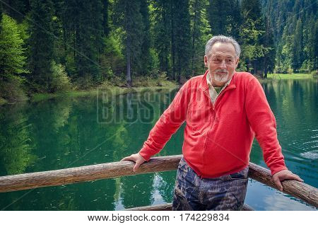 Portrait of a handsome senior man in bright sweater near the beautiful forest lake. Looking aside. Smiling happy senior man with gray hair and beard on excursion. Outdoors. Horizontal image.