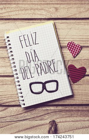 the text feliz dia del padre, happy fathers day in spanish written in the page of a notebook, a pair of black eyeglasses and some red hearts on a rustic wooden background
