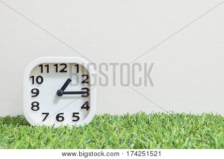 Closeup white clock for decorate show a quarter past one or 1:15 p.m. on green artificial grass floor and cream wallpaper textured background with copy space