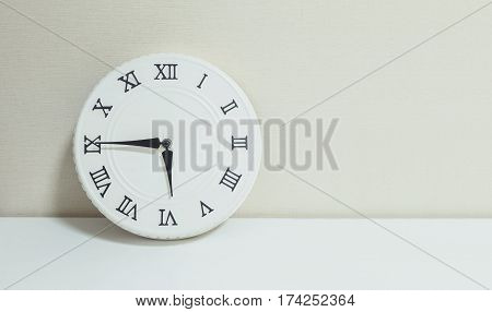 Closeup white clock for decorate show a quarter to six p.m. or 5:45 p.m. on white wood desk and cream wallpaper textured background with copy space