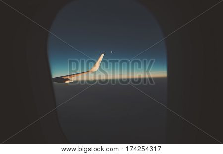 Contrast view through airplane porthole of horizon moon and dark teal morning sky illuminator of airplane with landscape outside dawn and part of the wing