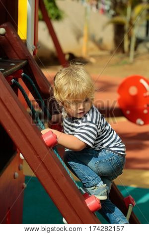 Cute baby boy with blond hair ponytail in blue tshirt and jeans climbing rope net or ladder on sunny summer day on playground background