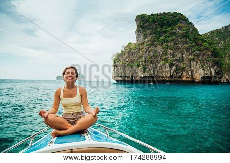 Beautiful girl with short hair in t-shirt and chert is sitting in a lotus position meditating on the deck of the yacht. Concept of calm and meditation.