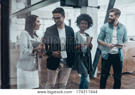 Working beyond the boarders of a desk. Group of young people in smart casual wear talking and smiling while walking together behind the glass wall in the creative office