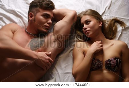 Young handsome athletic tattooed man lying in bed with gorgeous blonde in bicini passionately looking at each other shot from above
