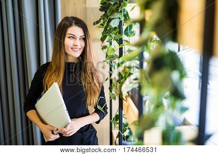 Portrait Of Successful Smiling Businesswoman Holding Laptop Computer In Her Hands. Happy Beautiful Confident Woman Standing At Office Wall With Notebook. Business People.