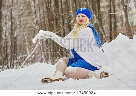 Plump smiling woman in skates sits in snowdrift and shakes off snow from mittens at outdoor skating rink in winter park.