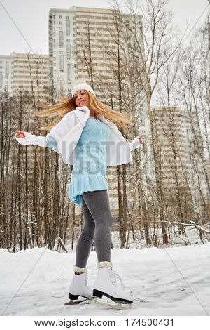 Young woman skates at outdoor skate rink in winter park against highrise buildings.