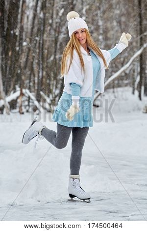 Young woman in blue knitted dress, white fur mantle and hat at outdoor skate rink on skates in winter park.